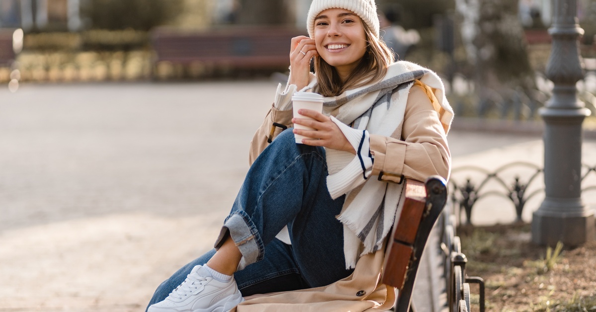A person sitting on a public park bench while drinking a hot drink. They are wearing a beanie, scarf, and long coat. A person sitting on a public park bench while drinking a hot drink. They are wearing a beanie, scarf, and long coat.