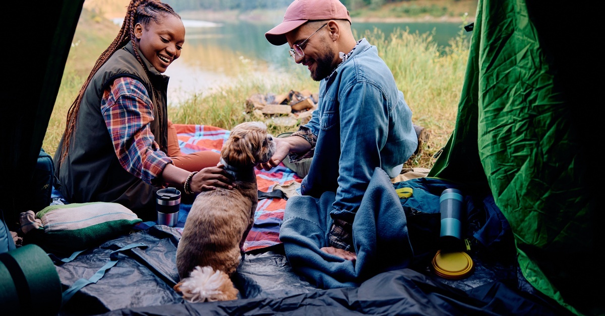 A pair of smiling people in cold-weather attire sitting in their camping tent alongside their small fluffy dog.