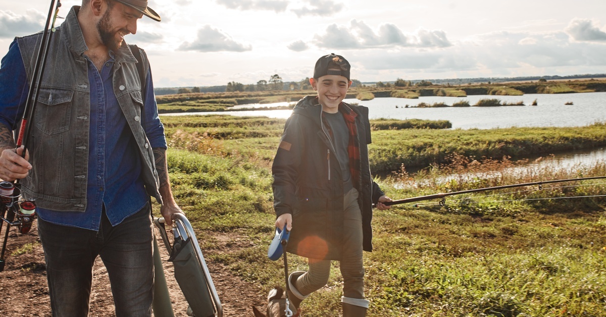 A father and son walking along the side of a marsh with their dog while carrying fishing gear, including rods and tackle.