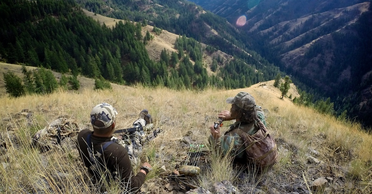 A pair of hunters resting at the top of a ridge in a mountainous area with many steep valleys on either side of them.