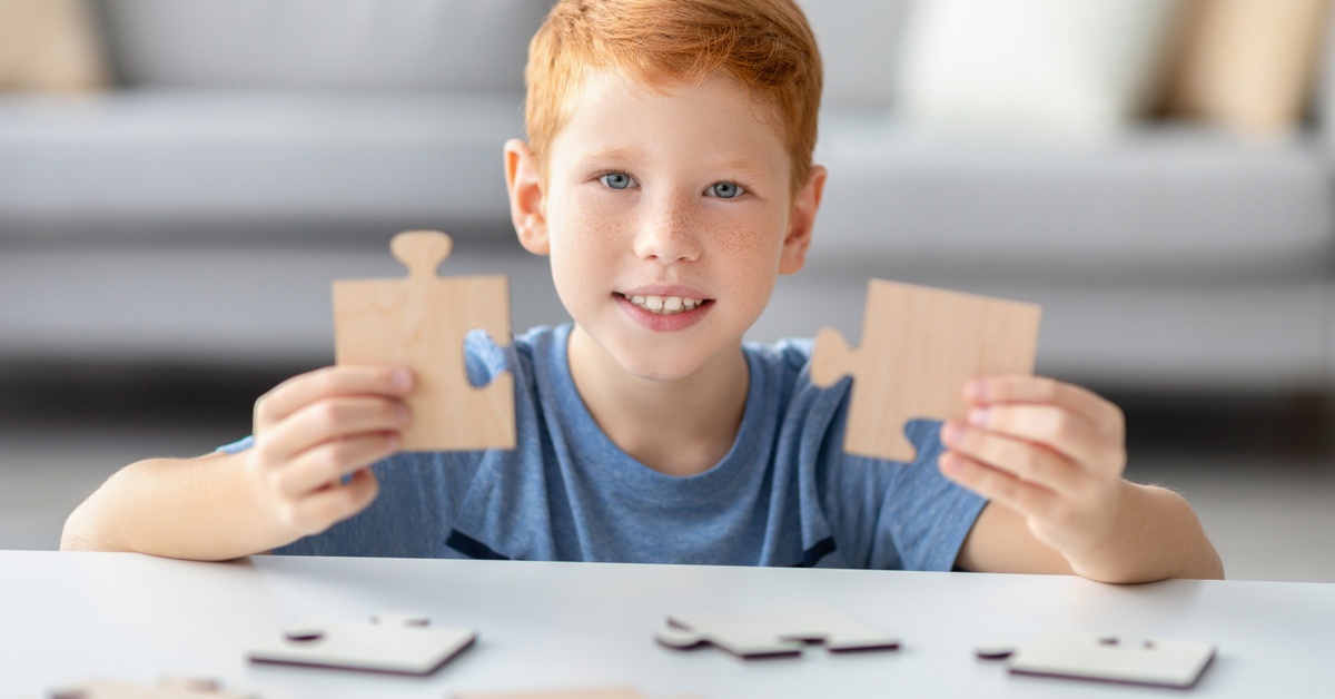 A child with bright hair sitting at a table covered in large jigsaw puzzle pieces. The child holds up two pieces and smiles.