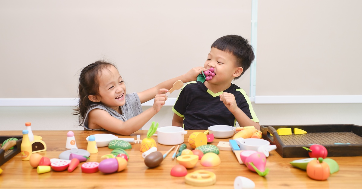 Two young children sitting at a table and playing together with toys in a pretend cooking set that resemble food.