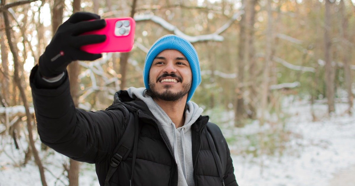 A young man standing on a snowy path and taking a selfie. He is wearing casual cold-weather attire to keep warm.