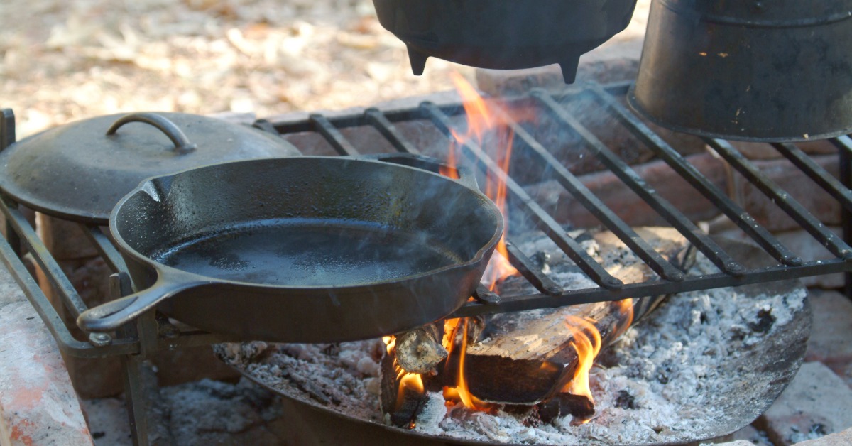 How To Season Cast Iron Properly for Nonstick Cooking A group of cast iron cookware sitting on an open grate above burning wood logs, including a cast iron skillet.