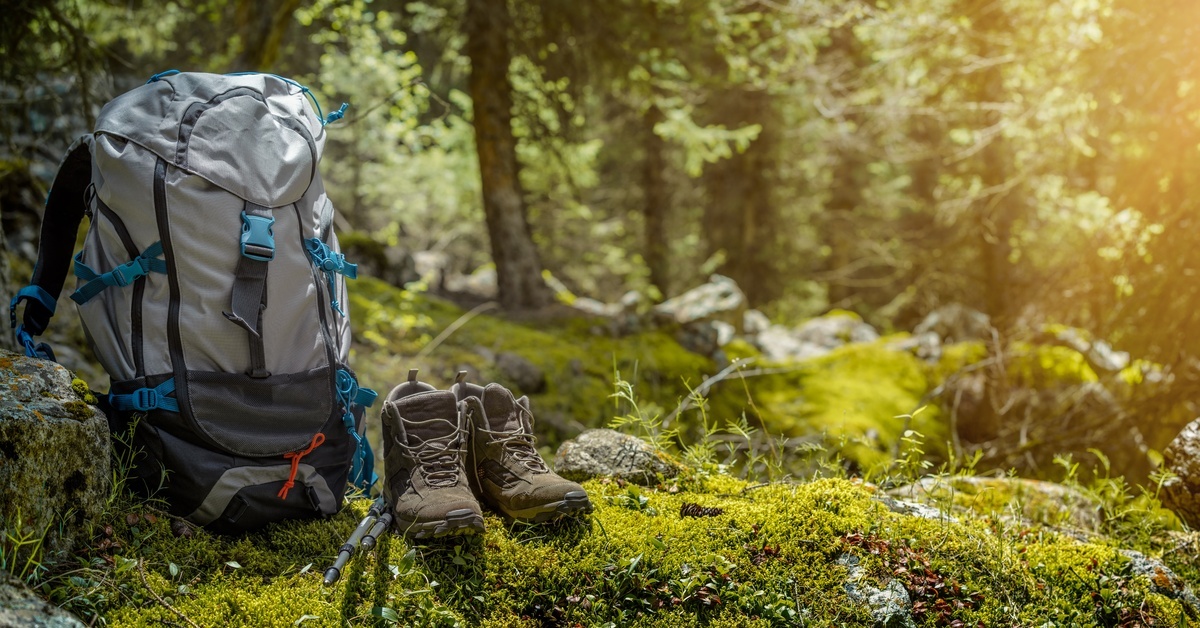 A light gray hiking or camping backpack sitting on a mossy rock beside a pair of clean brown hiking boots.