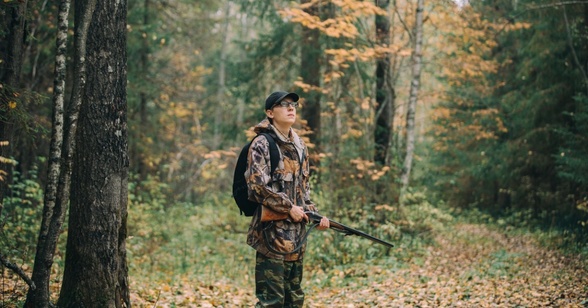 A hunter in camouflage print standing on a forested hill among deep green shrubbery and dark trees.