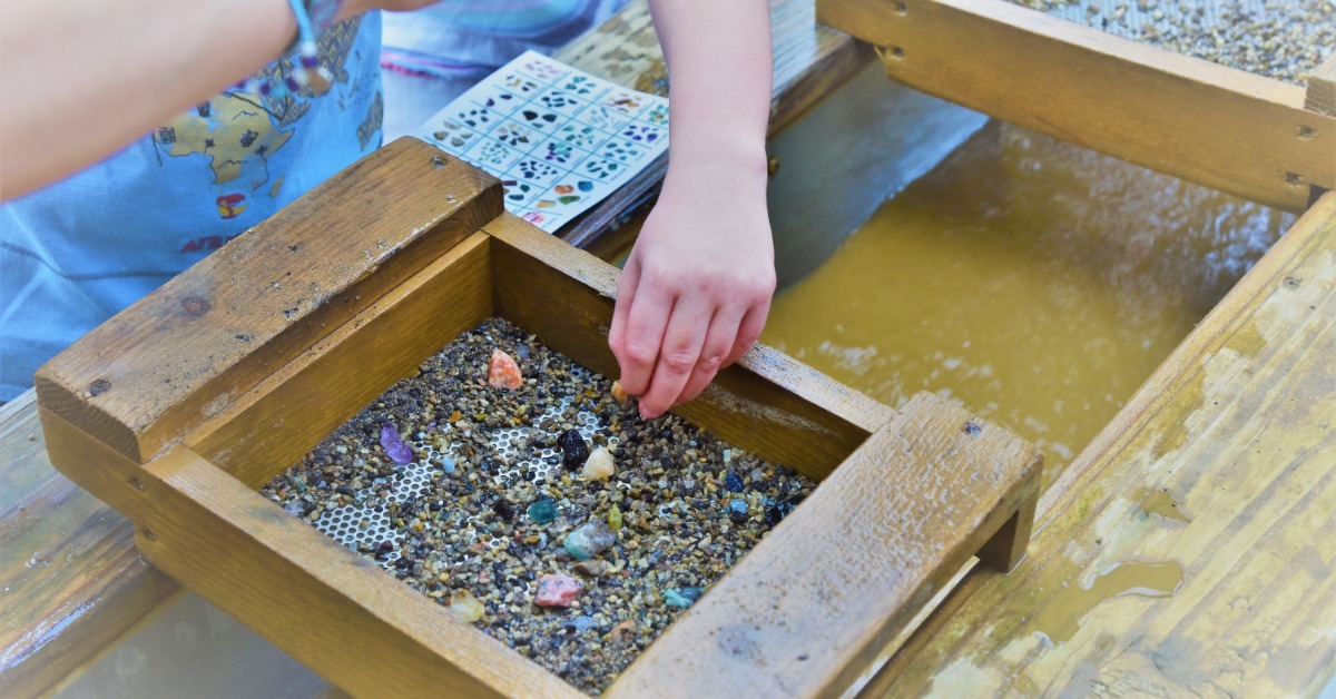 A group using wooden sluice boxes to separate colorful gemstones from small pebbles within an educational setup.