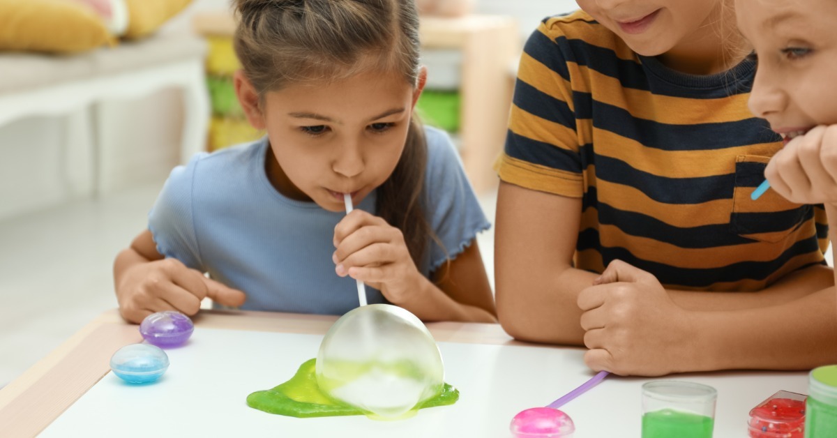 A group of children playing with colorful slime on a white table. One child blows an air bubble into a green slime pile.