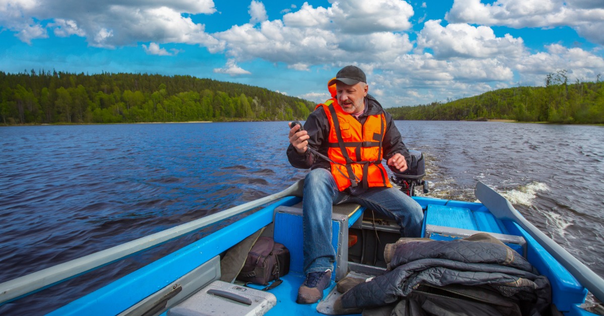 A man sitting on a blue-and-gray motor boat with gray oars on each side. He is setting up to fish while wearing layers.