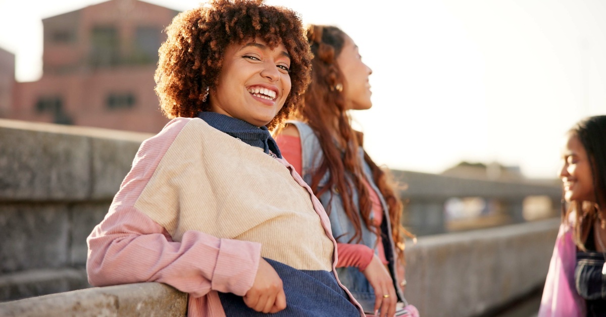 A smiling woman in a jacket with long sleeves leaning against a rail while standing with friends outside.