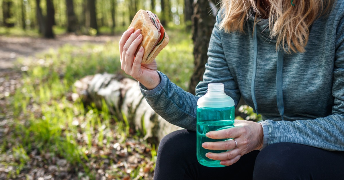 A person sitting down on a log while on a hike. They are holding a small sandwich and a plastic water bottle.