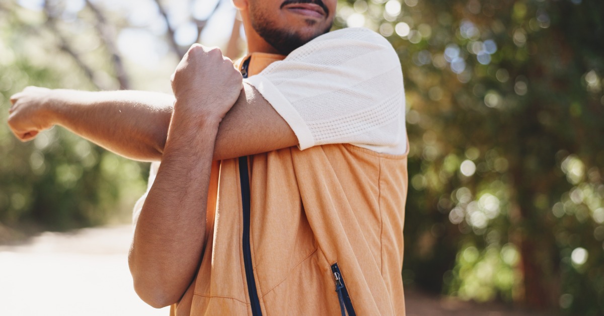 A person stretching while on a hike. They are wearing a simple lightweight jacket over a white top.