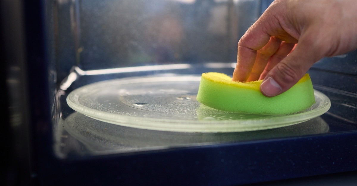 Close-up view of a hand using a yellow sponge to wipe moisture from the internal rotating plate of a black microwave.