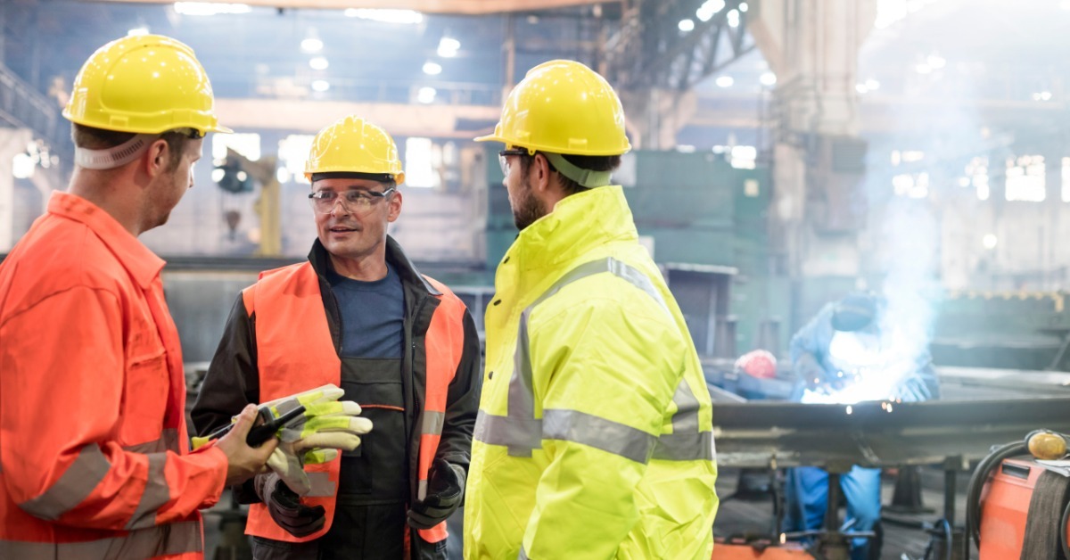 Three manufacturing workers standing inside a floor area while another worker welds metal in the background.