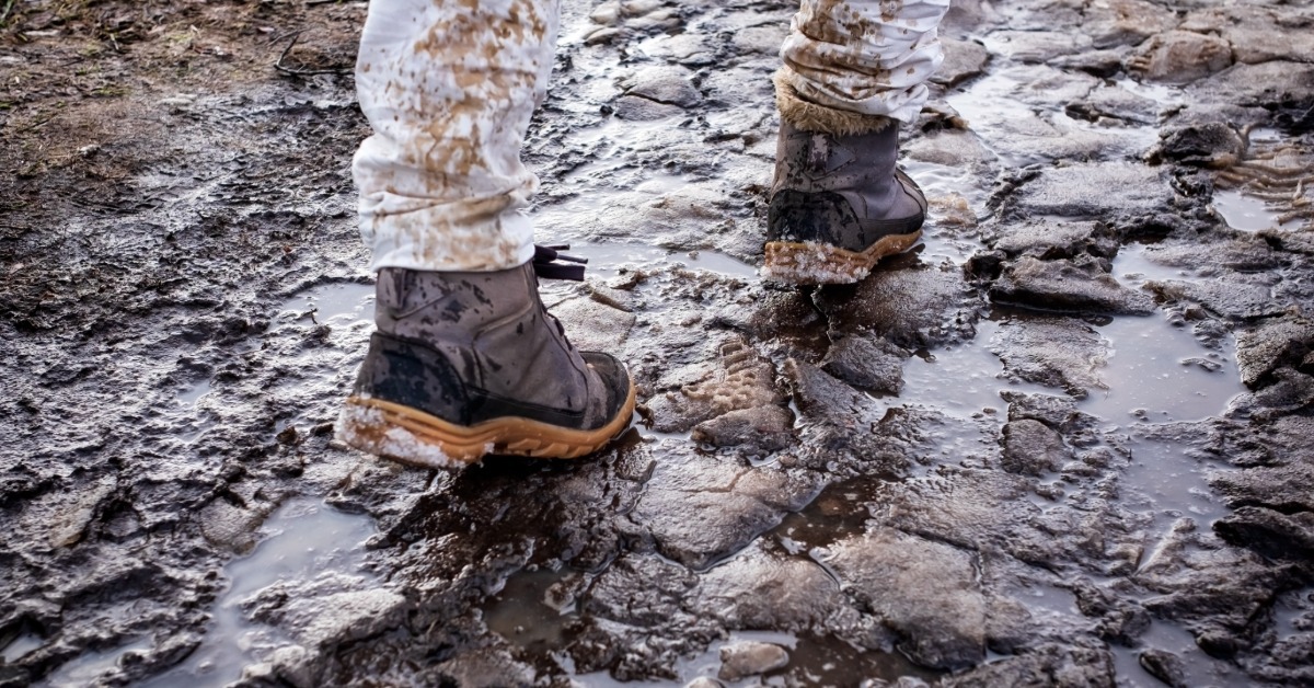 A person splashing in muddy water on an outdoor trail while wearing wet-weather boots and jeans.