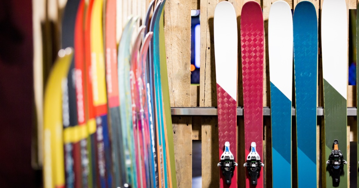 A collection of skis positioned upright in matching pairs against a wooden fence in bright sunlight.