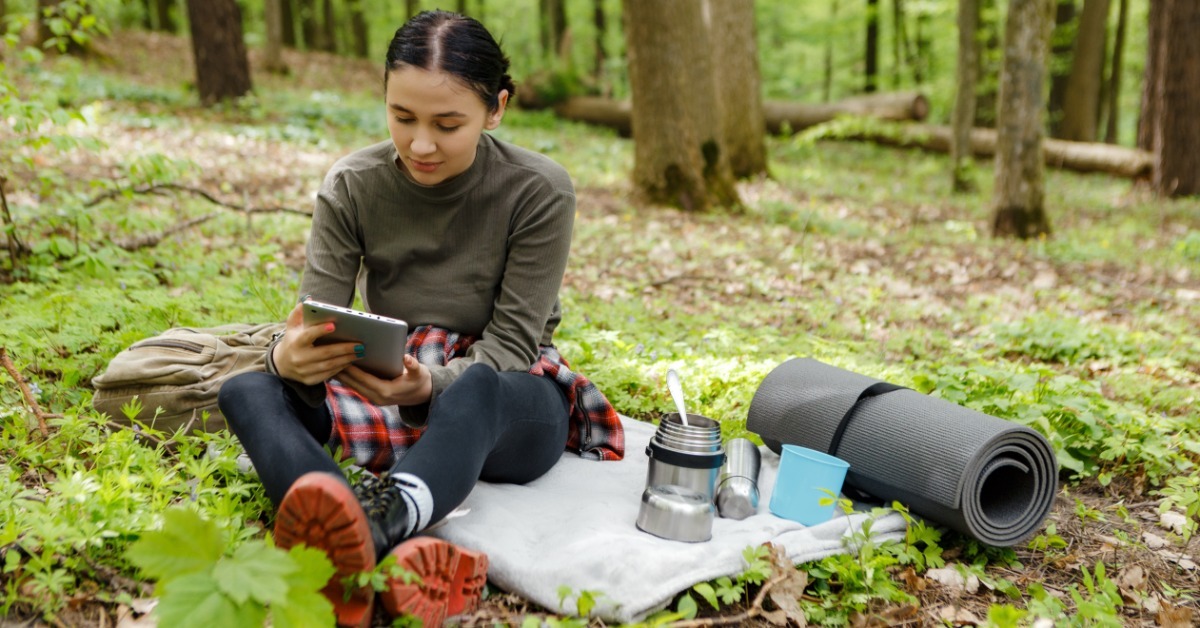 A hiker sitting on a cloth on the ground and reviewing information on a tablet. They have a mat and an open thermos.