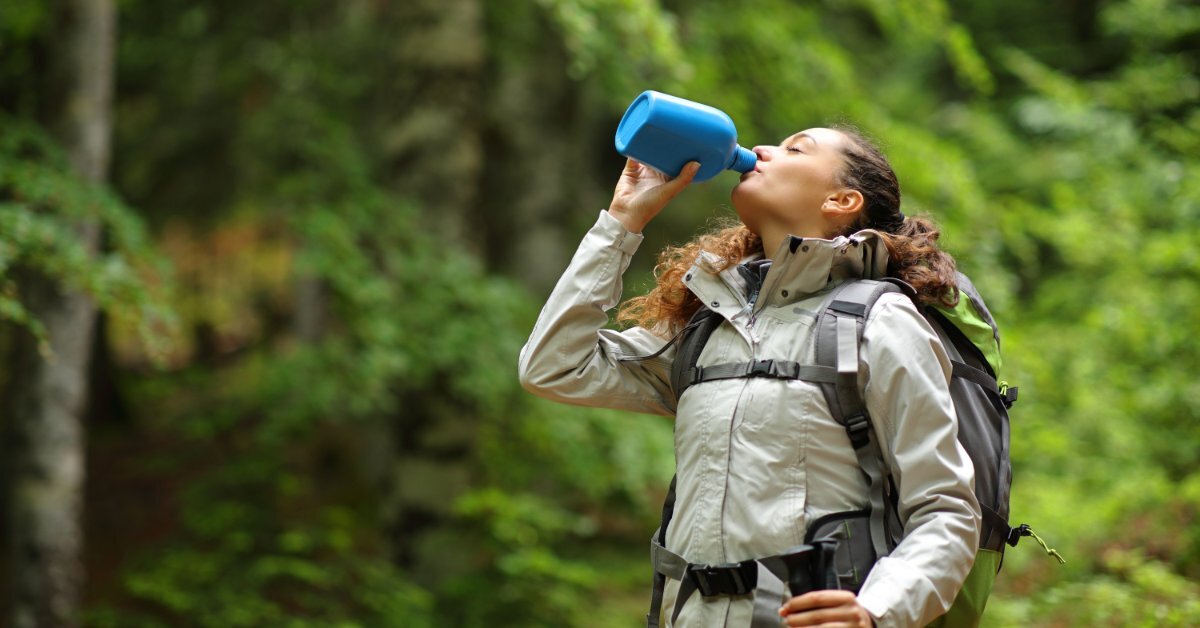 An outdoors enthusiast wearing camping gear and walking in a wooded area as they drink from a canteen.