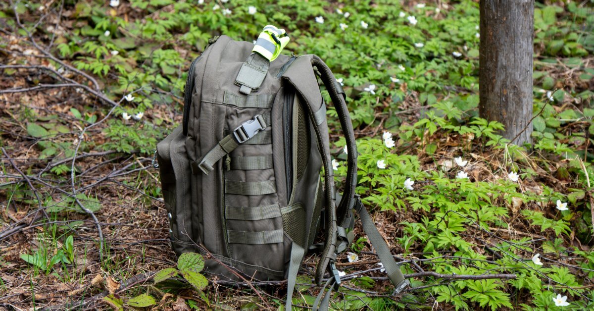 A tactical hunting backpack in a deep, natural green color sitting on a patch of plants and twigs in a section of forest.