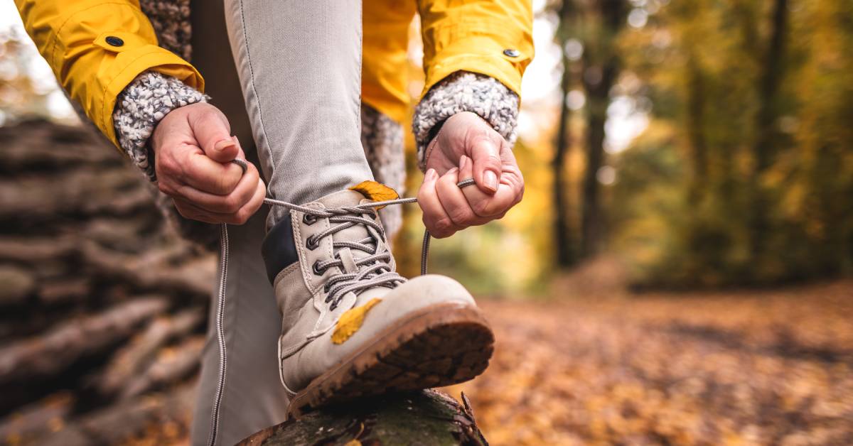 A close-up of a woman tying her hiking boots on a log in the forest. The boots and laces are a grayish-tan.