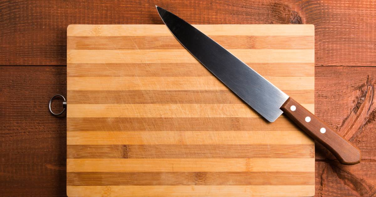 A large kitchen knife with a wood handle rests on a wooden cutting board over a dark wood kitchen table.