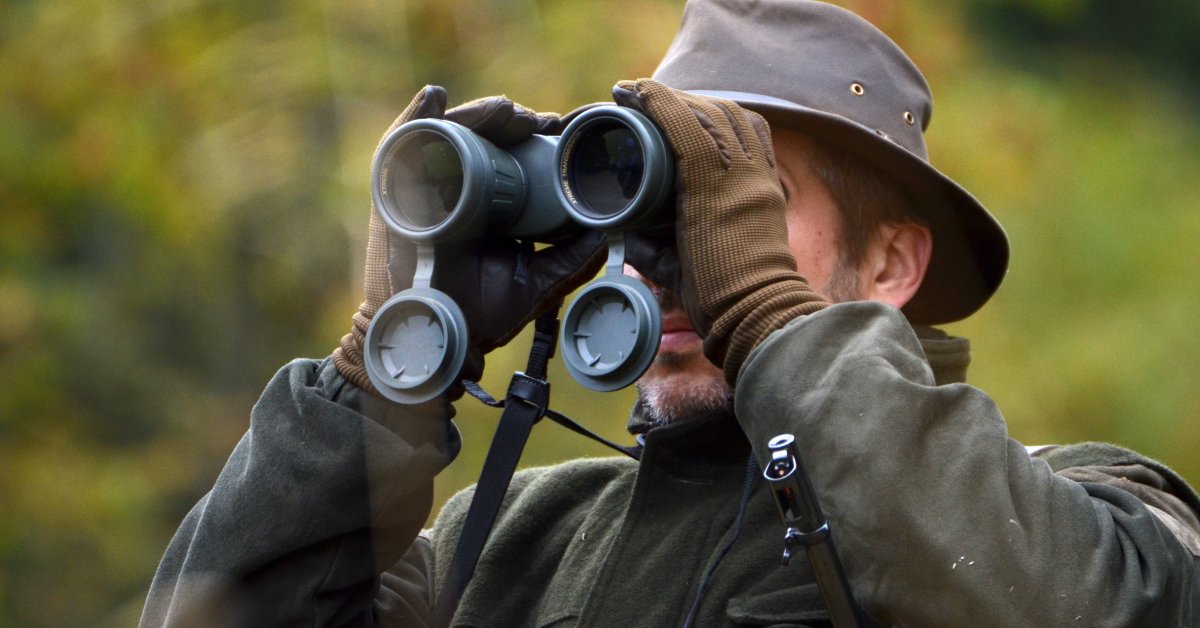 A hunter in focus wearing a green coat, brown gloves, and brown hat peers through binoculars against a blurred background.