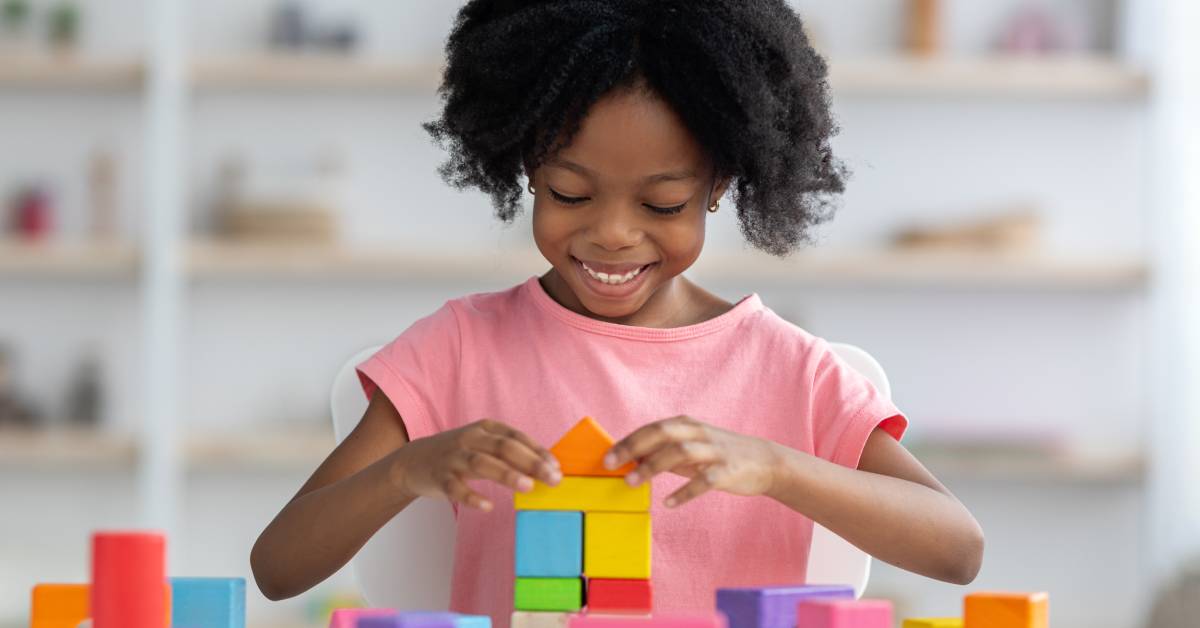 A young girl places an orange triangular building block toy on a structure of other various building blocks.