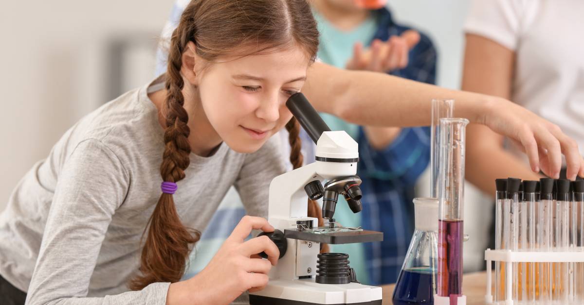 A young girl peers into a small microscope on a desk with various beakers and liquids surrounding her.