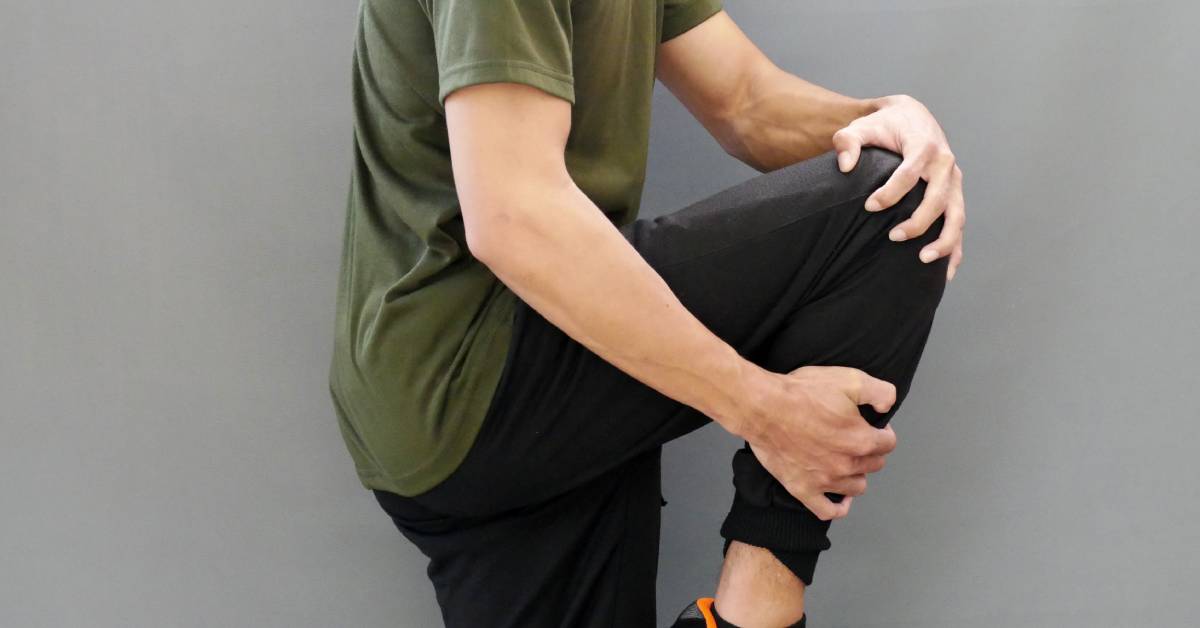 A man in a green t-shirt and black jogger pants with black and orange sneakers stretches his legs against a gray background.