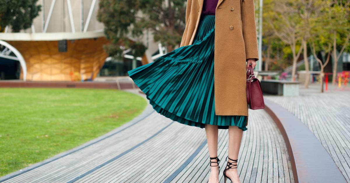 A woman in a red sweater and green pleated midi skirt with a beige overcoat and red handbag poses outside.