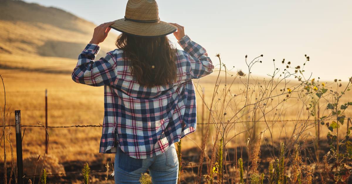 A woman on a farm walks away from the viewer in a flannel top, jeans, and straw hat with a rural landscape behind her.