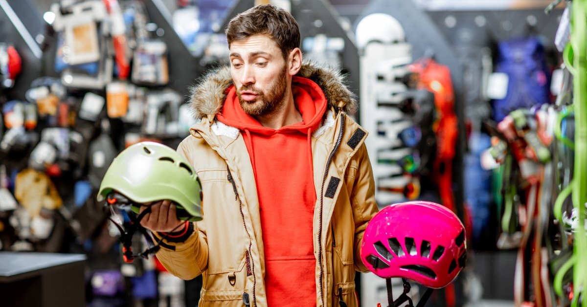 A man in a winter coat and red hoodie looks at mountaineer helmets, one green and one pink, in a sporting goods store.