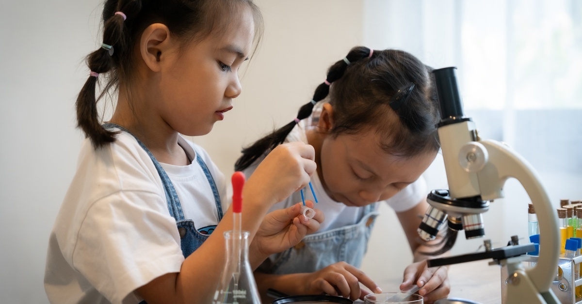 Two young girls in matching outfits play with a kid's chemistry see,t including a microscope and beakers.