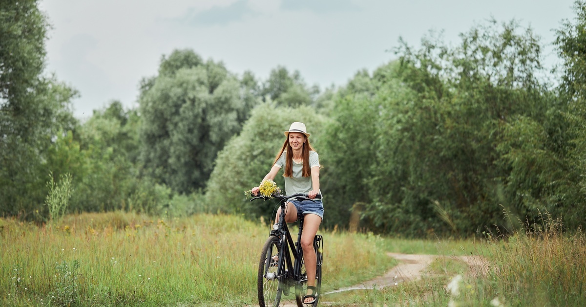 A young woman in a green shirt and fedora rides on a bike with flowers on the front on an outdoor trail.