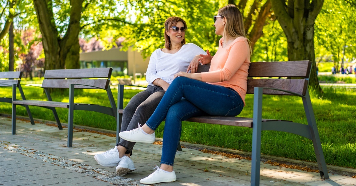Two adult women, dressed casually in jeans and colorful tops with sunglasses, talk on a bench in a park.