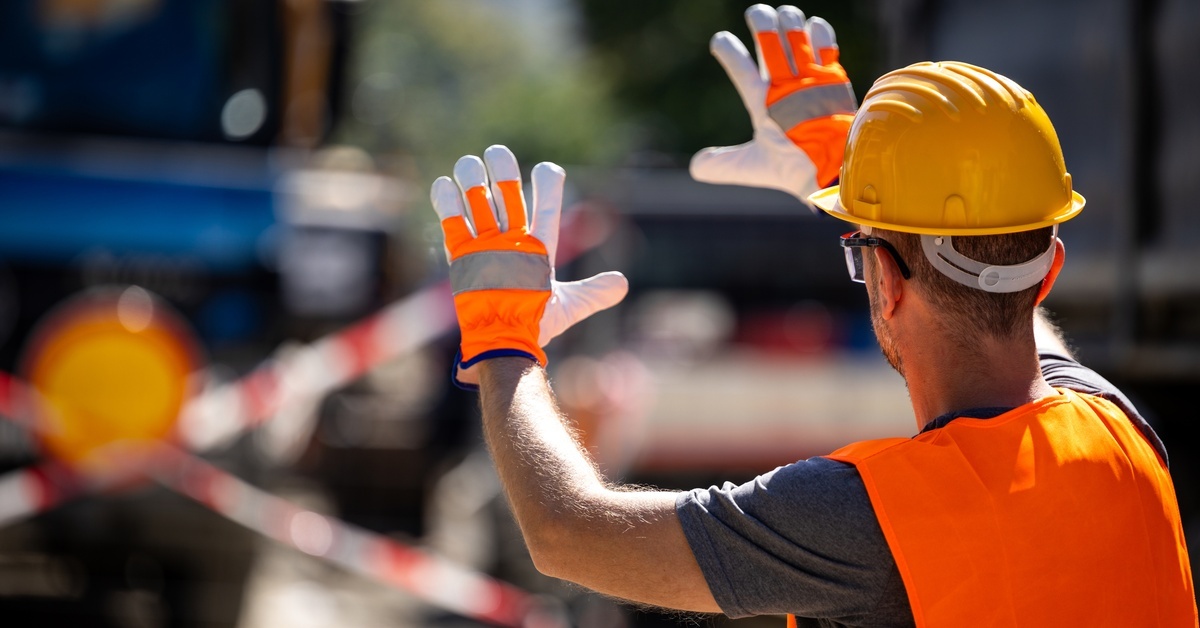 A construction worker raising his hands in front of a vehicle. He is wearing a bright pair of orange gloves on his hands.