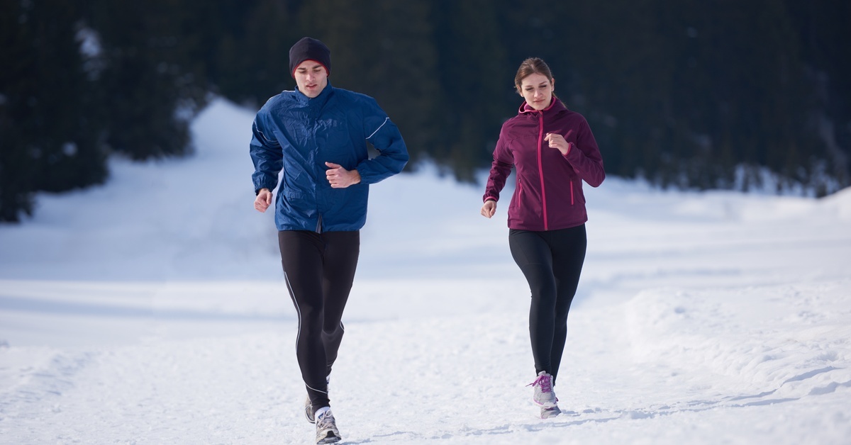Two people are jogging on packed snow. One person wears a blue jacket, the other wears a plum-colored jacket.