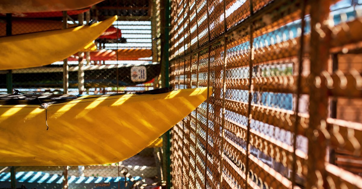 Four kayaks are stacked inside a covered area. There's a window covered with blinds, letting in beams of light.