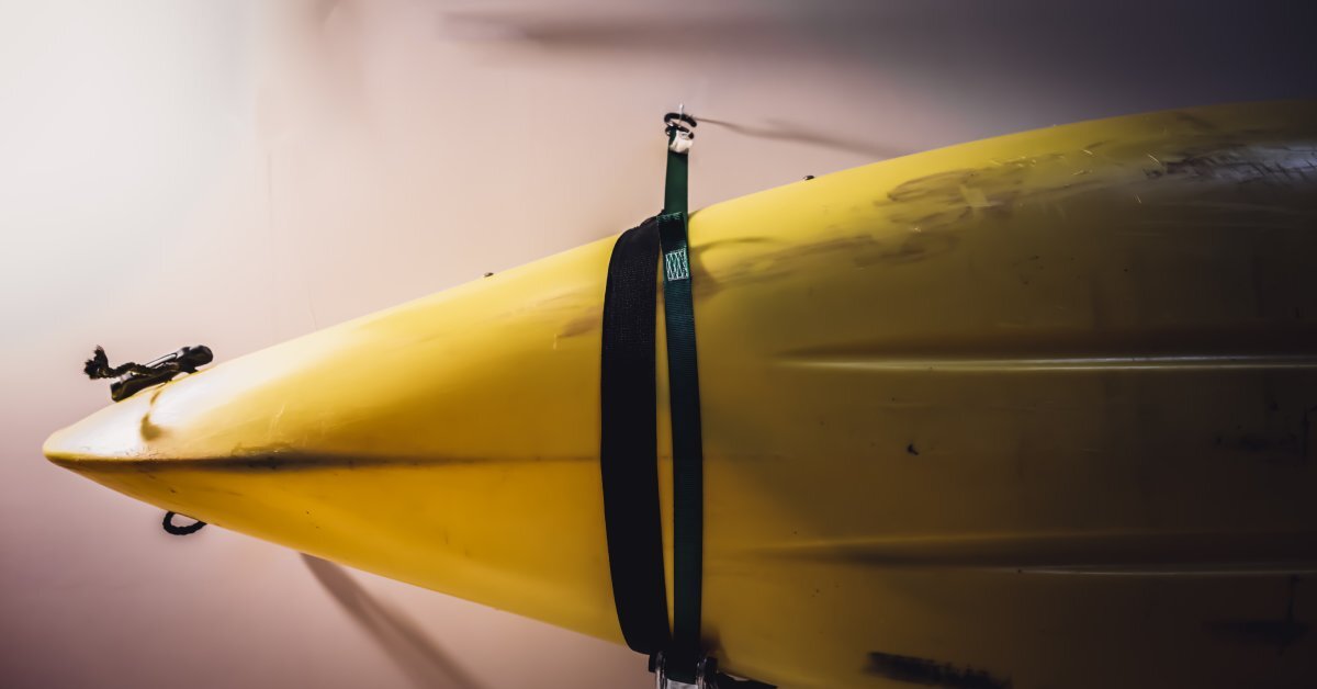 A close-up of a yellow kayak. The watercraft is suspended in the air with a black strap and is attached to a white wall.