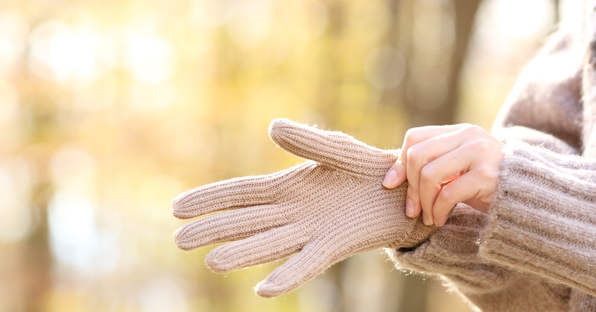 A person uses their bare hand to put a beige glove onto their other hand. The background is full of trees and blurred. A person uses their bare hand to put a beige glove onto their other hand. The background is full of trees and blurred.