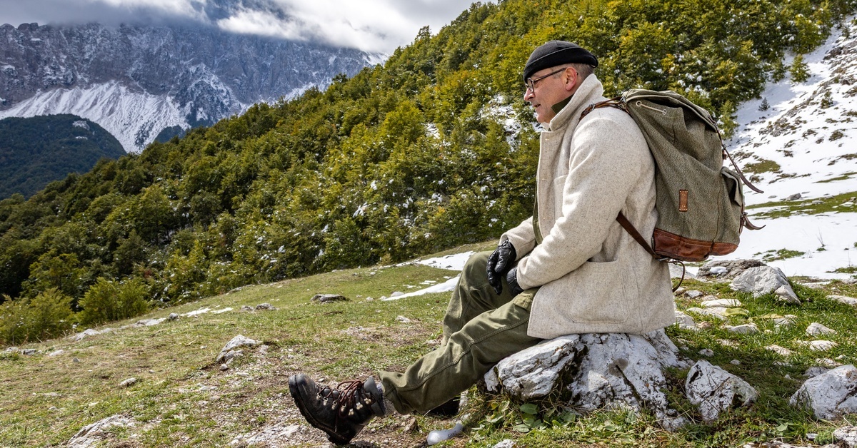 A man wears a coat, boots, and a backpack as he sits on a rock. Green hills and snow-covered mountains are in the distance. A man wears a coat, boots, and a backpack as he sits on a rock. Green hills and snow-covered mountains are in the distance.