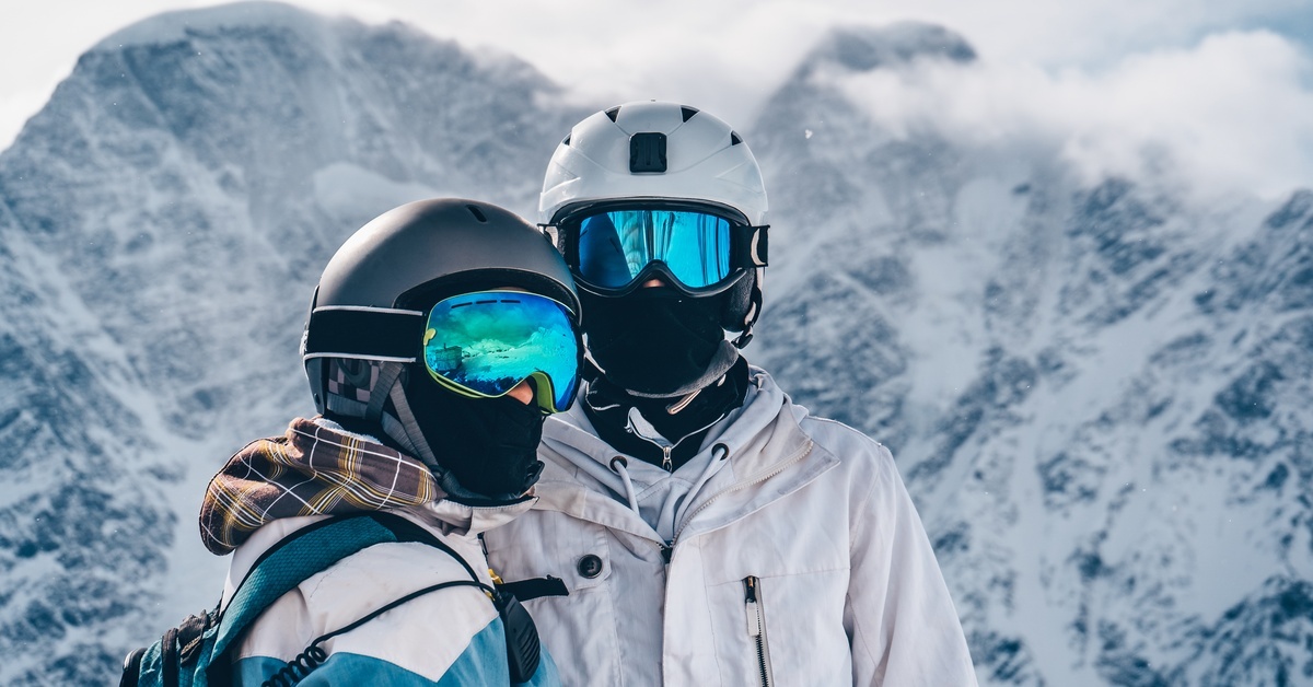 Two people wearing coats, blue ski goggles, and helmets are standing in front of a snowy mountain range.
