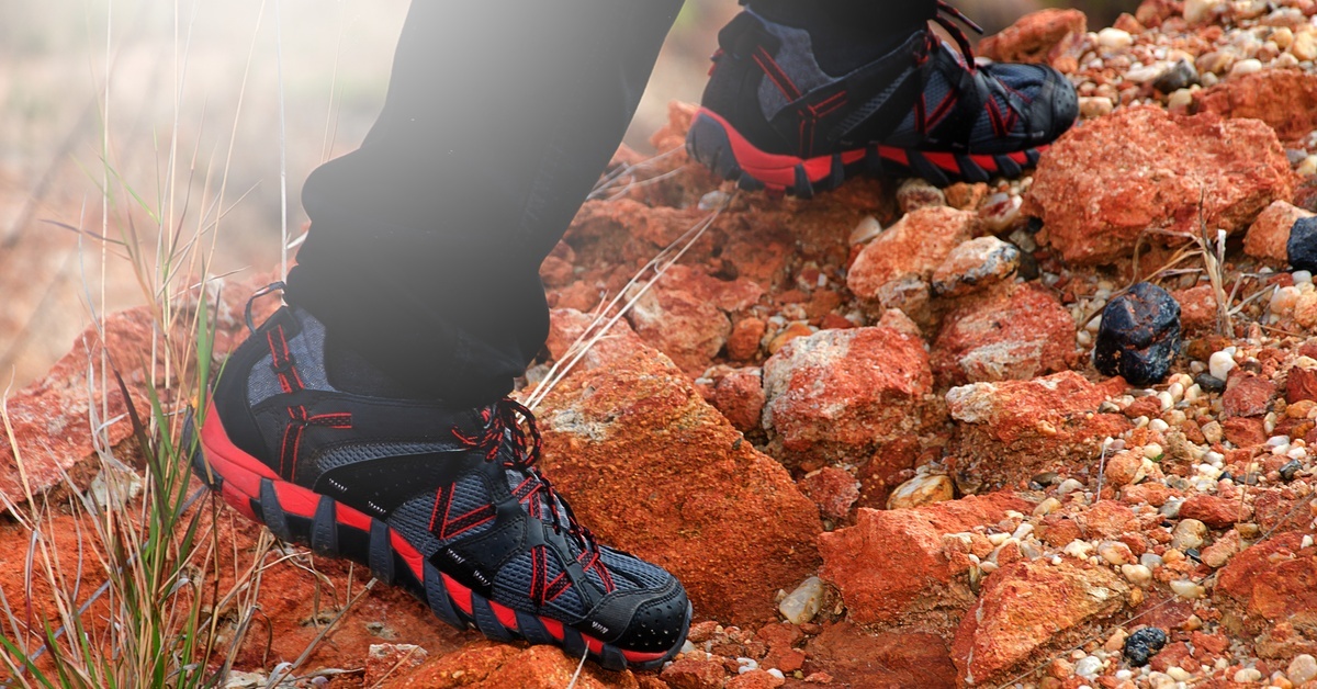 A person wearing red and black sneakers and black pants stands on orange-red rocks. The sun shines in the distance.