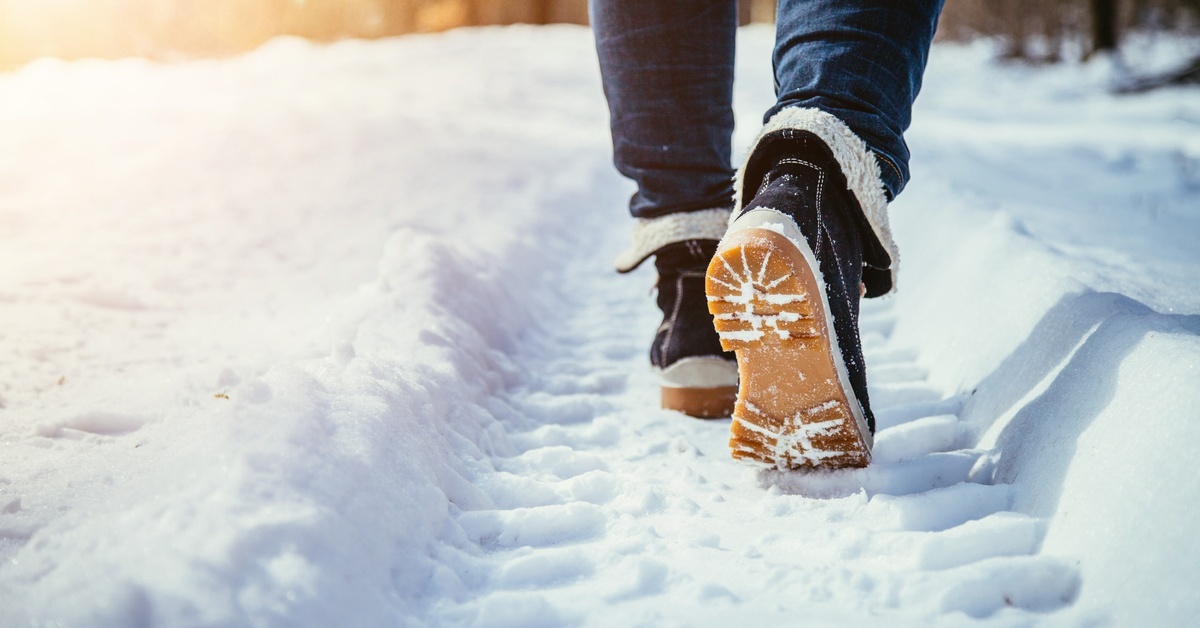 A close-up of a person walking in packed snow from a large tire. The shoes are black with a tan sole.