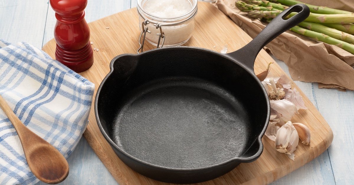 A cast-iron skillet, cloves of garlic, and a jar of salt sit on a wooden board beside a towel and a wooden spoon.