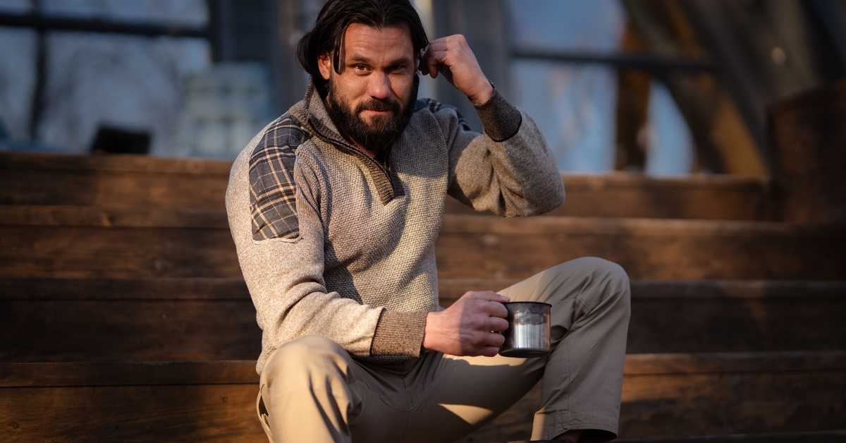 A man in an earth-tone outfit with a natural fabric sweater sitting on a wooden porch and drinking coffee.
