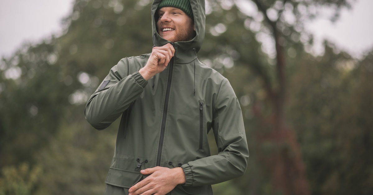 A man wearing a natural sage green rain shell jacket with the hood up and the jacket zipped while outdoors.