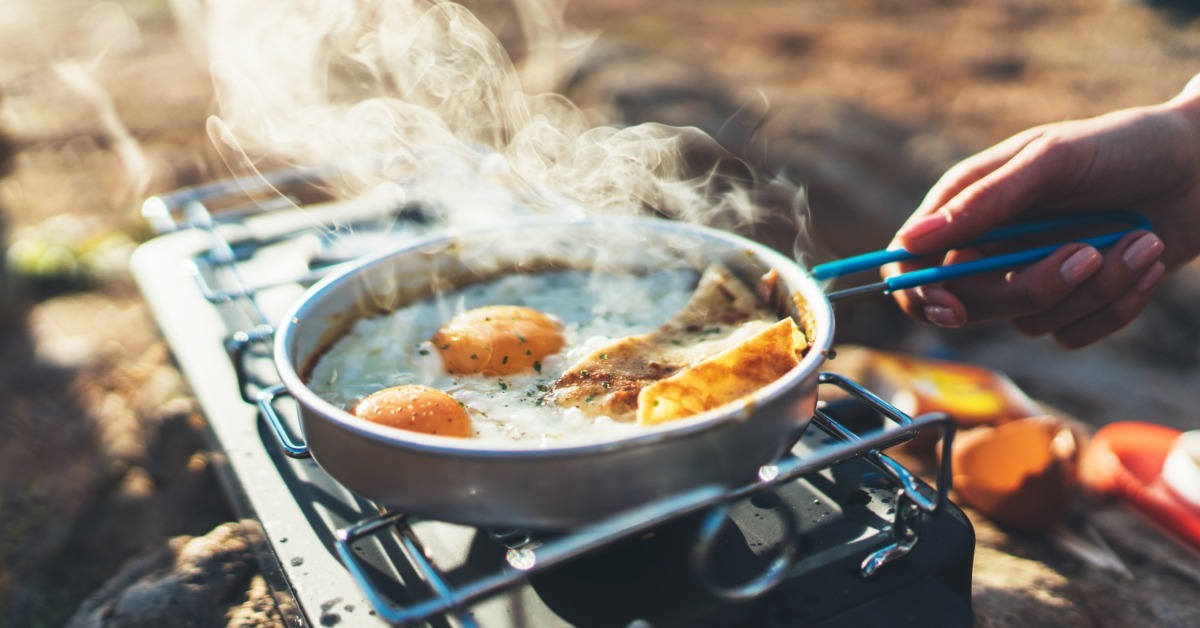 A hand holds a blue utensil over fried eggs cooking in a pan on a portable gas stove with steam rising.