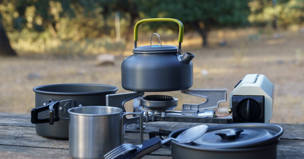 A gray kettle sits on a portable stove with pots, a mug, and utensils on a wooden table in front blurred trees.