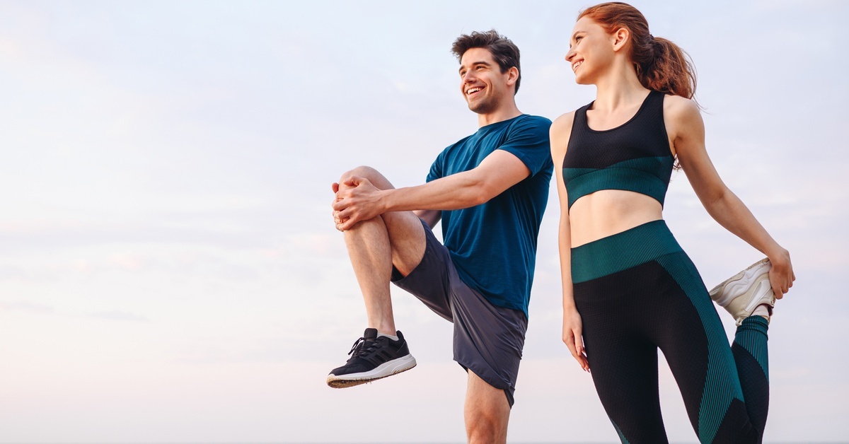 A man and woman wearing athletic clothes stretch together on a beach near calm water beneath a pale sky.